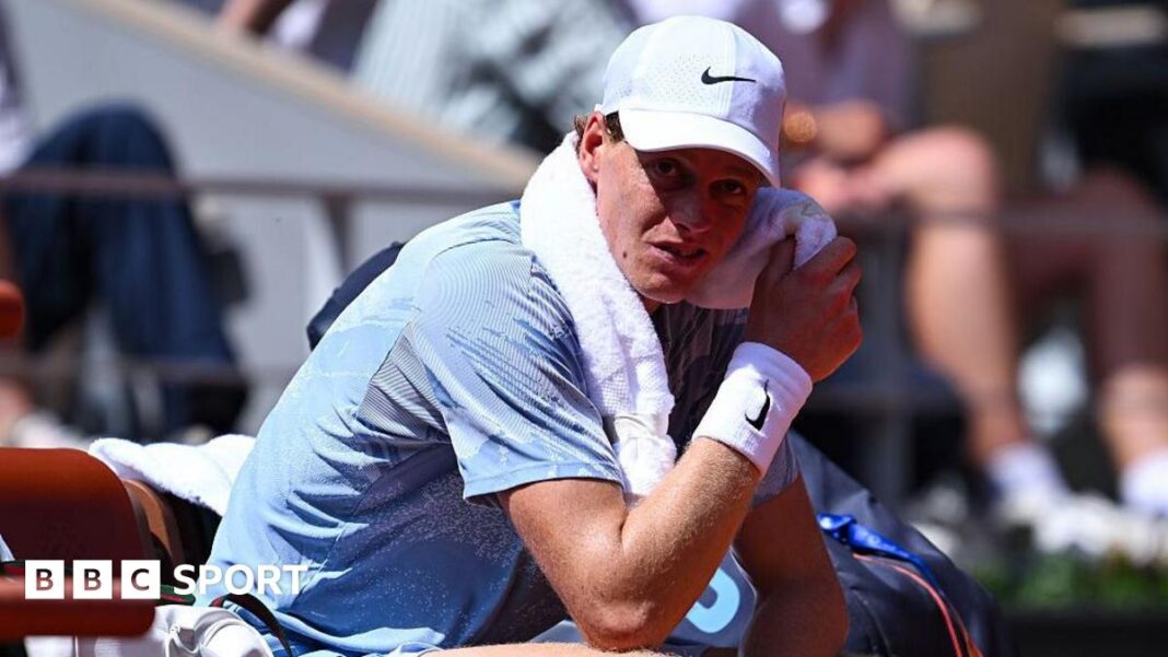 Jannik Sinner, wearing a grey t-shirt and white cap, sits in his chair with a towel around his neck wearing a pained expression